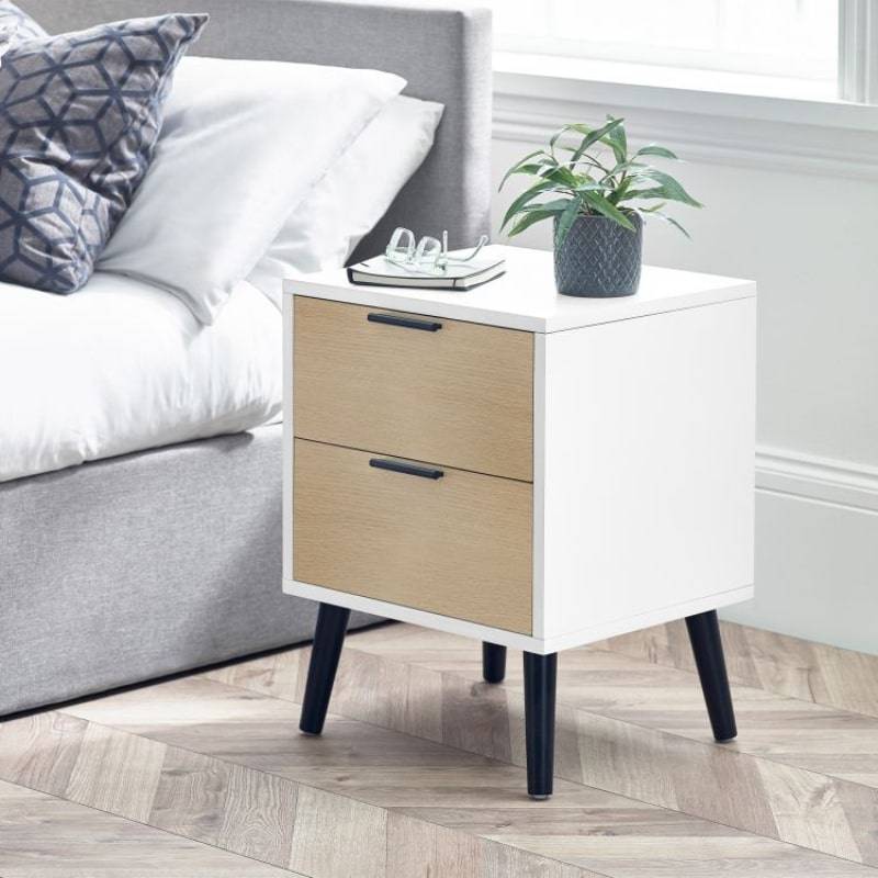 Alba bedside cabinet in white and oak effect finish with two drawers, black handles, and angled legs, styled next to a bed in a modern bedroom.