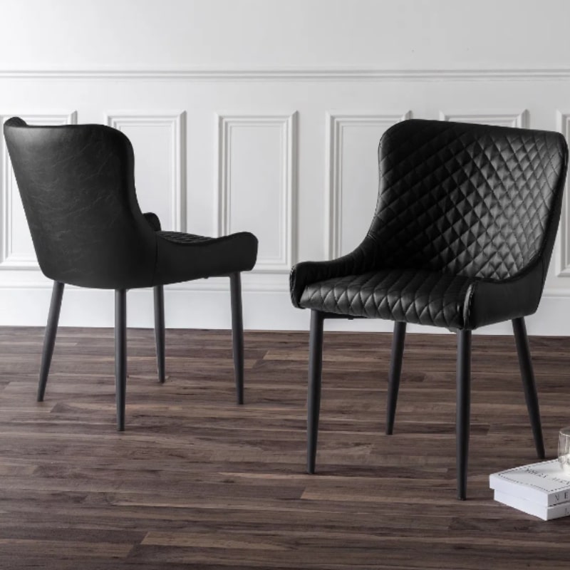 Set of two black Luxe Faux Leather Dining Chairs with quilted detailing and sleek black legs, placed in a modern dining room with wood flooring.